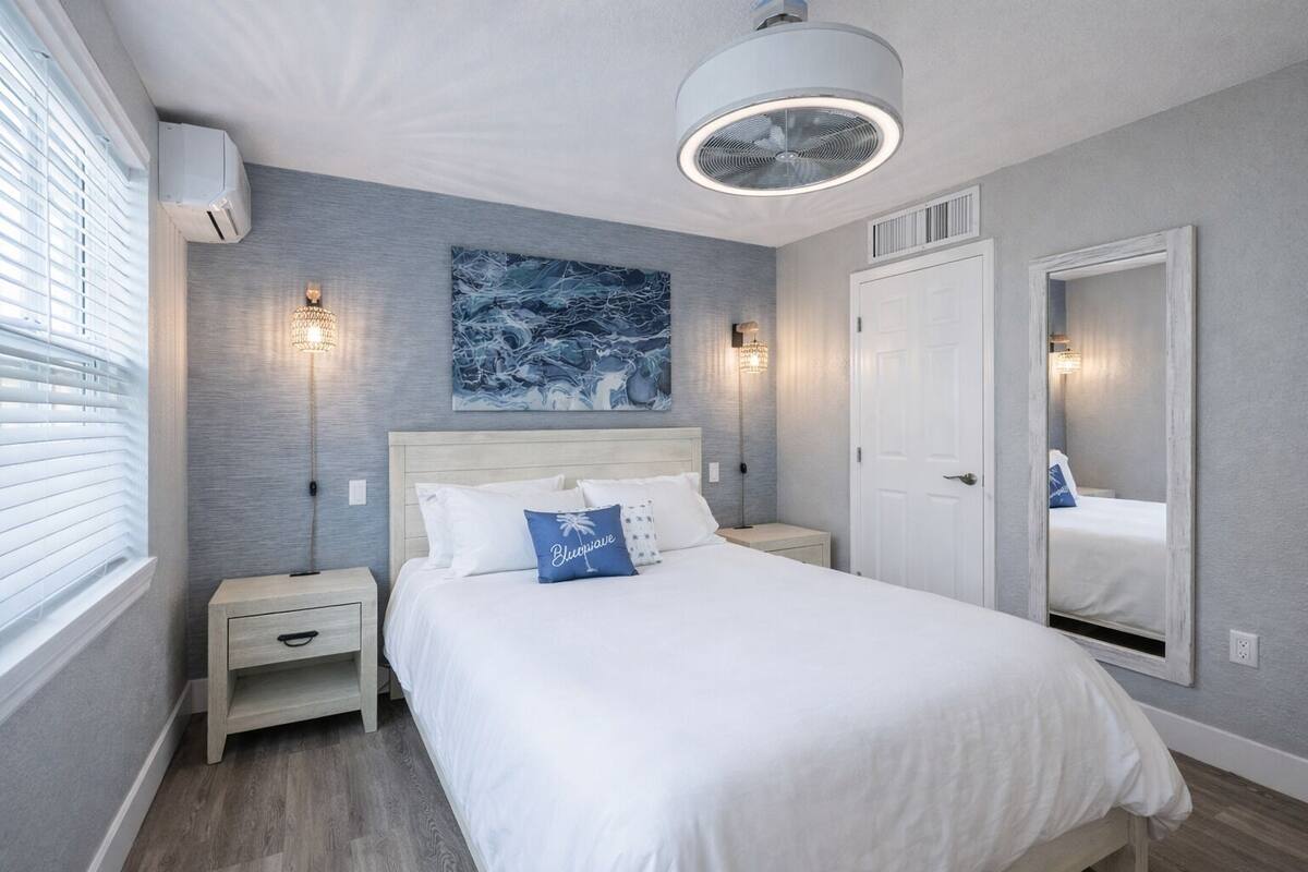 A bedroom with a bed, bedside tables, a ceiling fan, a large mirror, and artwork on the wall.