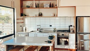 A modern kitchen with white cabinetry, a stainless steel refrigerator, and a built-in oven. There are bar stools at the island and a window providing natural light. - Kowhai Cabin - Serene Scandi Escape in Hokitika (Hokitika)