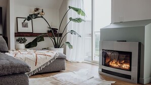 A modern living room with a fireplace, grey sofa, white rug, and large green plants.