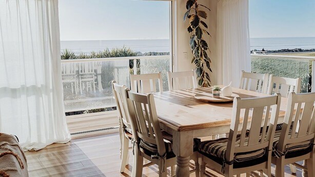 A dining area with a wooden table and white chairs, large windows with white curtains, and a view of the sea.