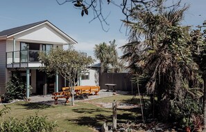 Una casa moderna de dos pisos con un balcón, un patio con mesa y sillas, y una variedad de plantas tropicales.