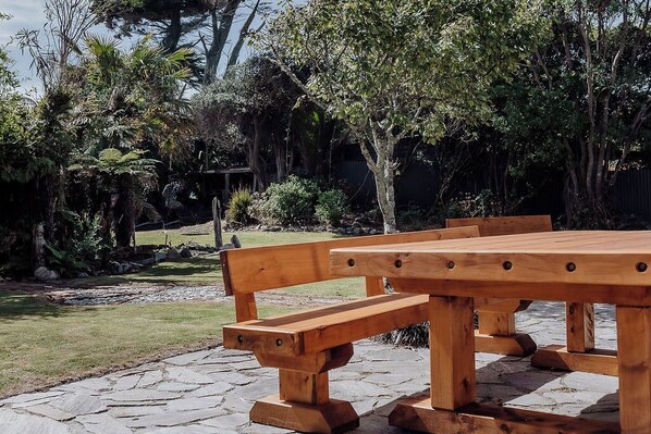 Un tavolo da picnic in legno con panche in un giardino con pavimento in pietra, circondato da diversi alberi e arbusti.