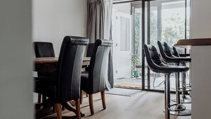 A dining area with a wooden table and four chairs, a bar area with stools, and a large sliding glass door leading to an outdoor space. - The Sundowner Beach Club (Hokitika)