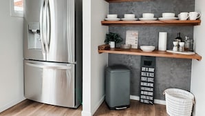Una cocina moderna con un refrigerador de acero inoxidable, repisas de madera con vajilla, un bote de basura y una cita motivacional en la pared.