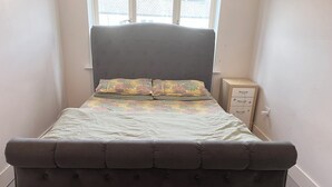 A bed with a floral bedspread and two pillows, a small bedside table with a lamp, and a window with blinds.