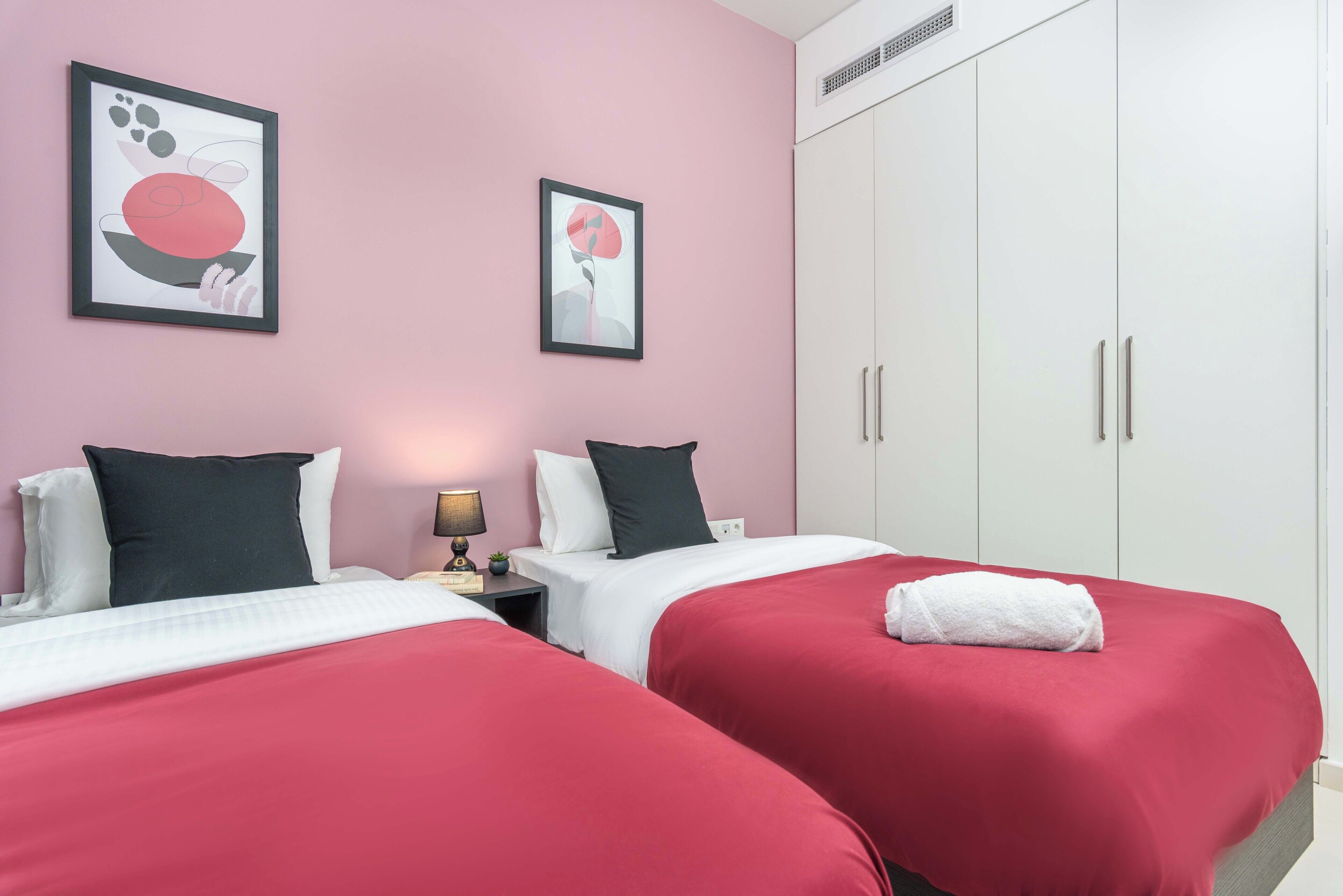 Habitación de hotel con dos camas, cada una con ropa de cama roja y almohadas blancas, una mesita de noche con lámpara y un armario grande.