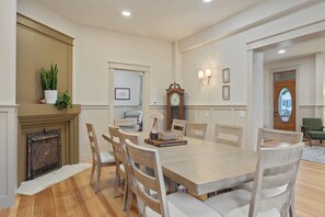 Une salle à manger avec une table en bois, six chaises, une cheminée, une horloge et des lampes fixées au mur.