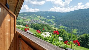 A wooden balcony with a view of a valley, green fields, and a town in the distance.