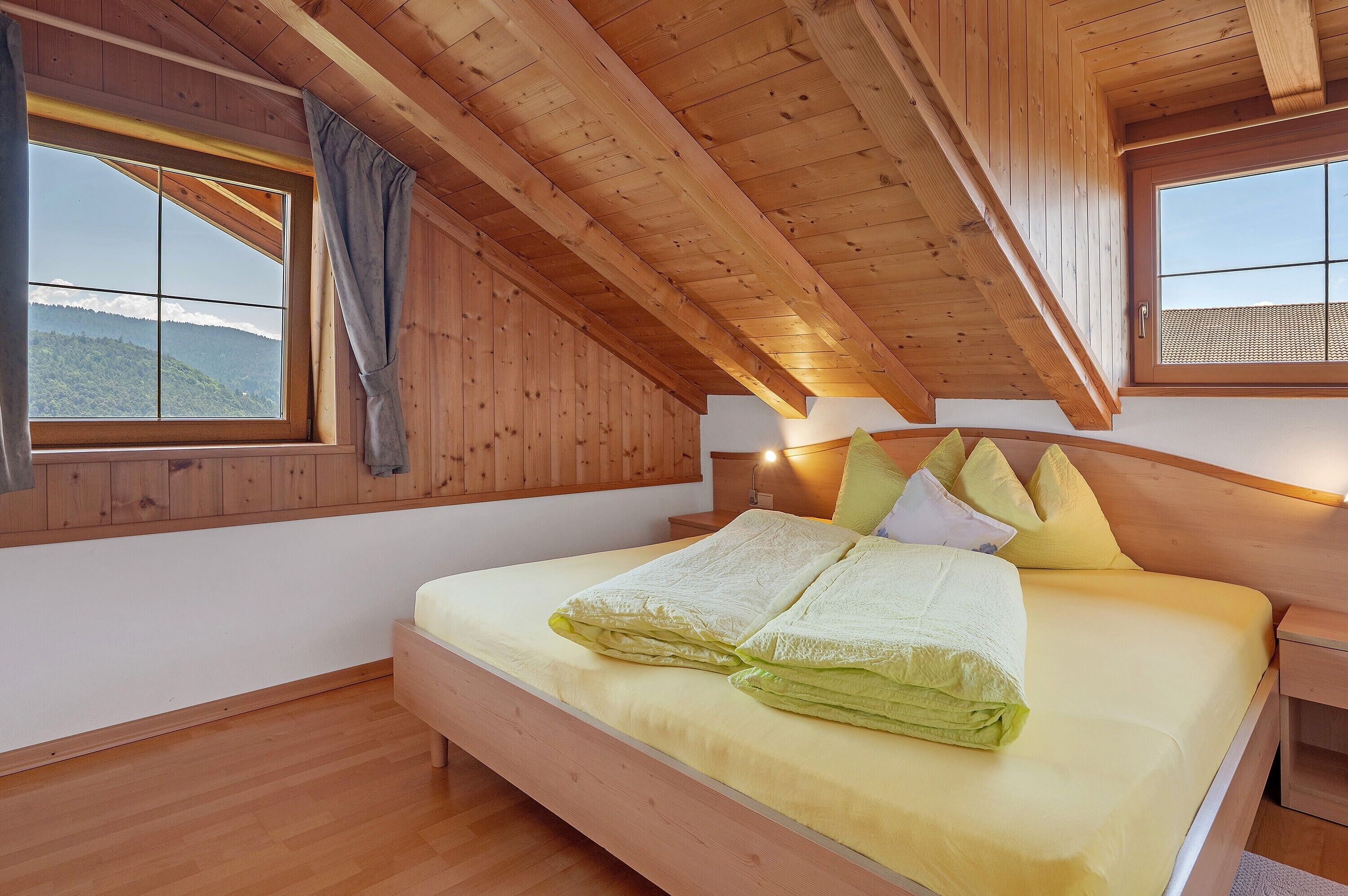Une chambre avec un plafond en bois, un lit avec une literie jaune, deux lampes de chevet et une fenêtre donnant sur les montagnes.