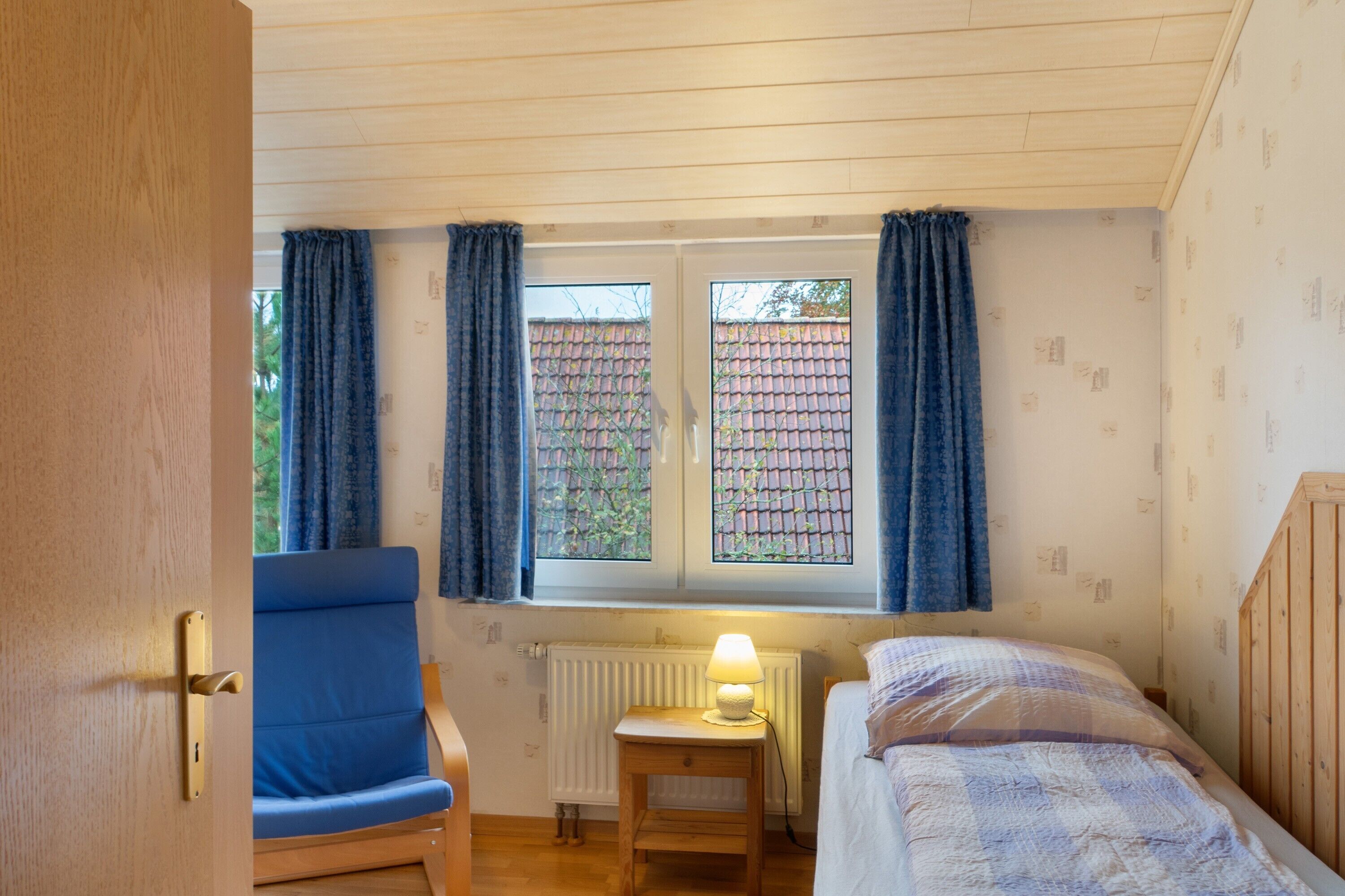 Ein Zimmer mit Holztür, blauem Stuhl, Nachttisch mit Lampe, Bett und einem Fenster mit Vorhängen.