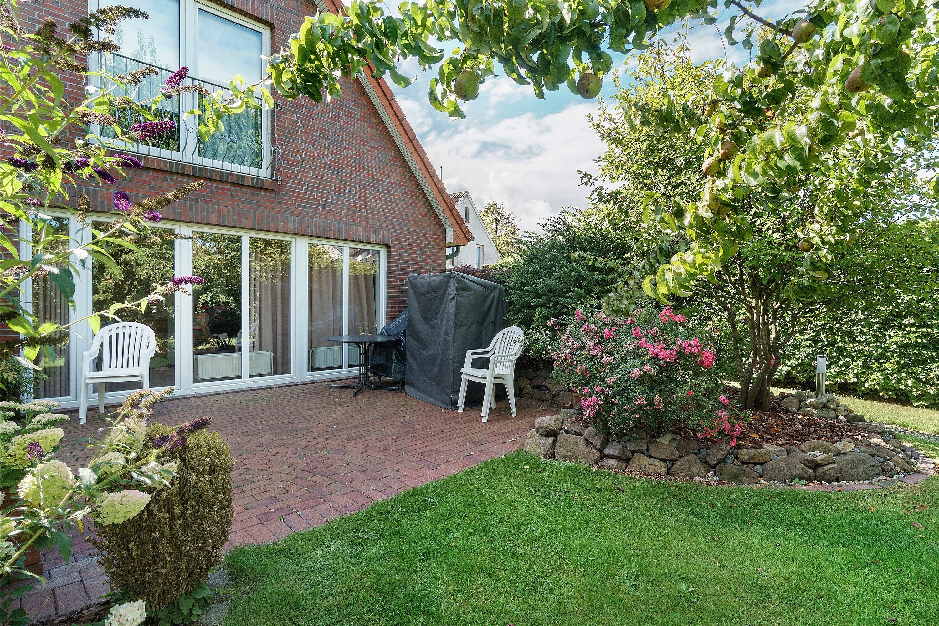 A house with a brick exterior, a covered patio area with two white chairs, and a garden with a tree and pink flowers.