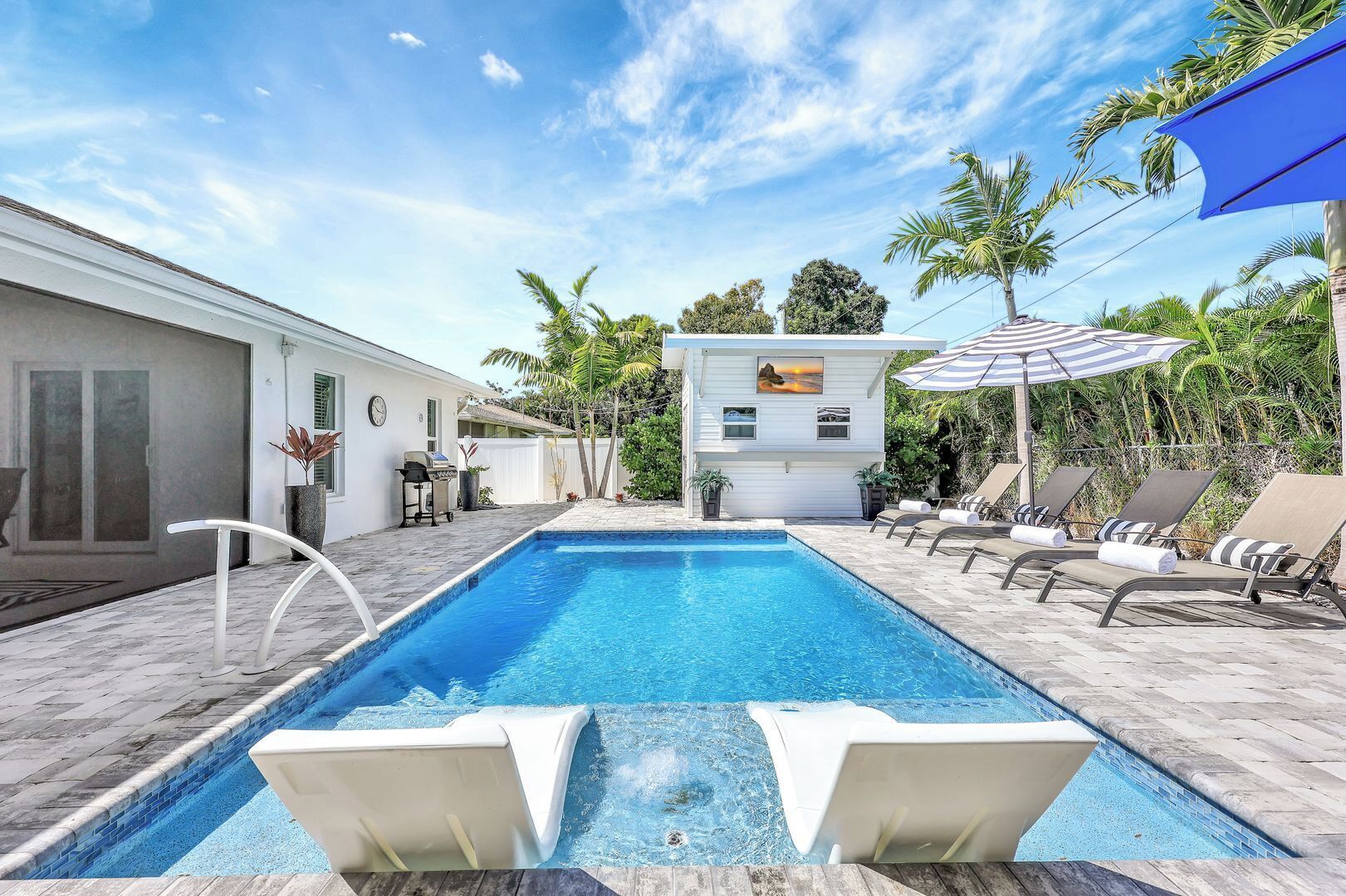 A rectangular swimming pool with two white lounge chairs in the foreground, a white building with a picture on the wall, and palm trees in the background.