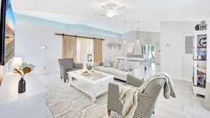 Een moderne woonkamer met een bank, salontafel en gestreept beklede stoelen. Er is een eethoek met tafel en stoelen, en een keuken is zichtbaar op de achtergrond.