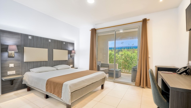 A modern hotel room with a large bed, a desk, and a balcony with a view of greenery.