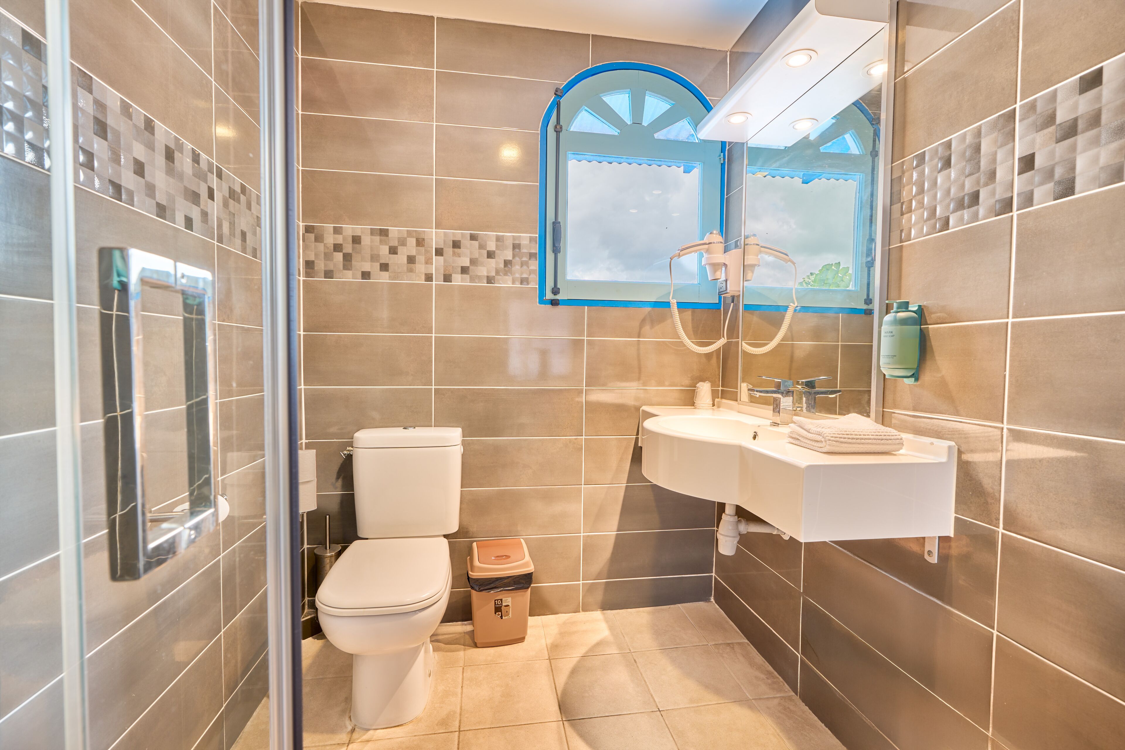 A modern bathroom with a glass shower enclosure, a toilet, a sink with a towel, and a mirror with a blue frame.