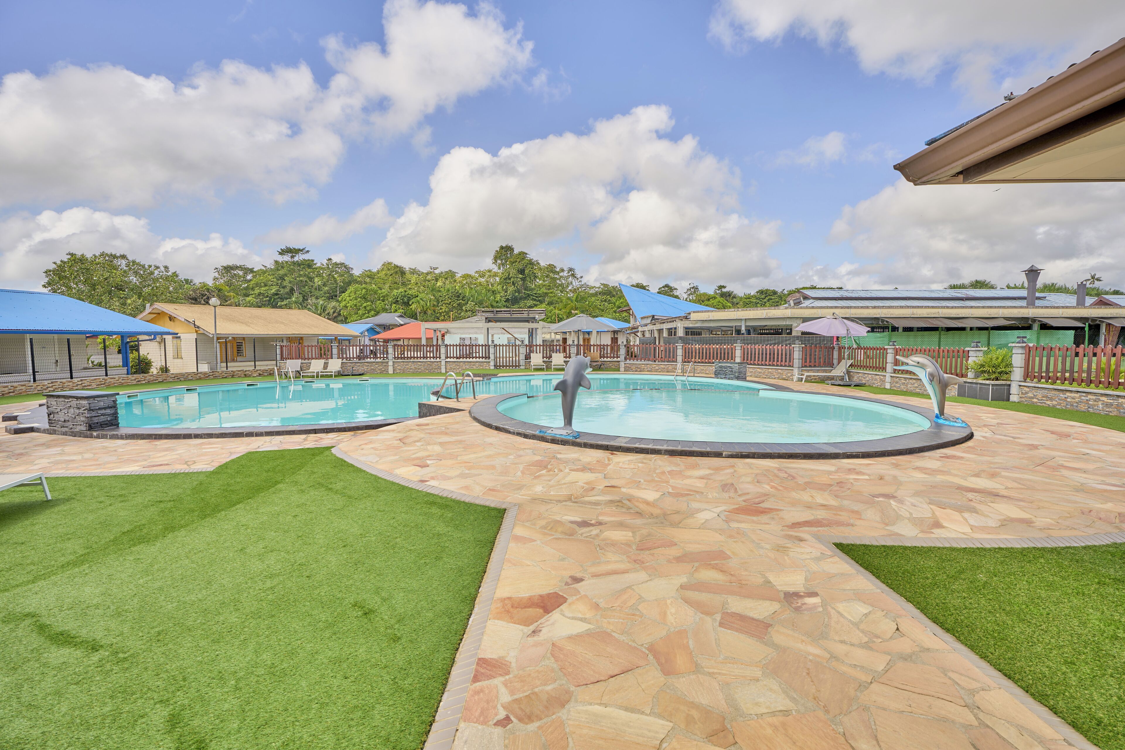 A swimming pool area with two pools, stone flooring, and green artificial grass.