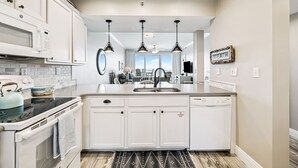 A modern kitchen with white cabinetry, a built-in microwave, a stove, and a dishwasher. There are pendant lights above the sink and a large window in the background. - HP 35W Gulf Front Condo @ High Pointe | Lagoon Pool | Tennis & Pickleball Courts (High Pointe Resort)