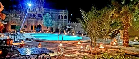 A nighttime outdoor pool area with tables and chairs, surrounded by palm trees and lit by lanterns.