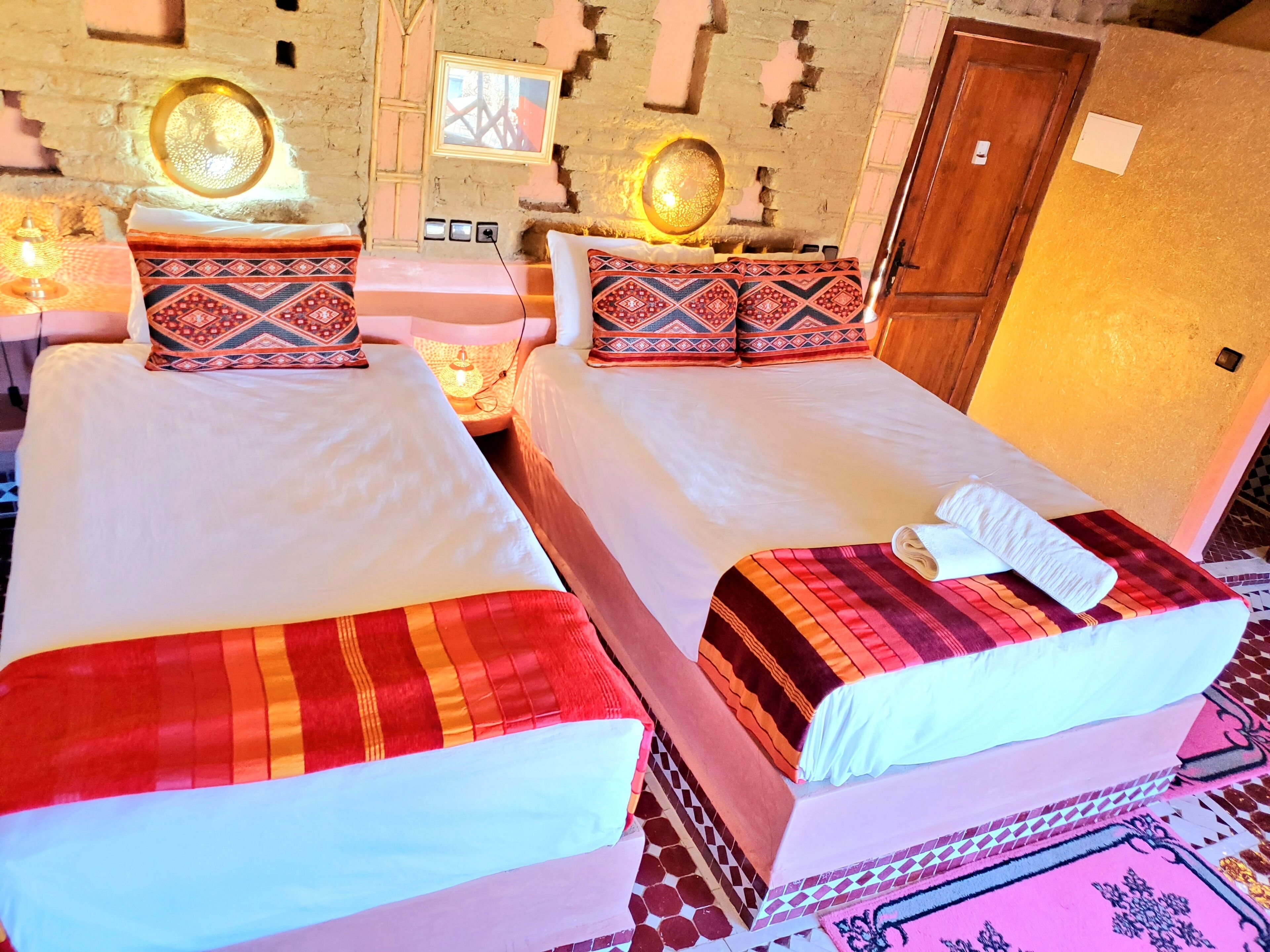 Ein Hotelzimmer mit zwei Betten, jedes mit gemusterten Kissen und einer roten und orangefarbenen Decke. An der Wand befinden sich Lampen, und es gibt eine Holztür.
