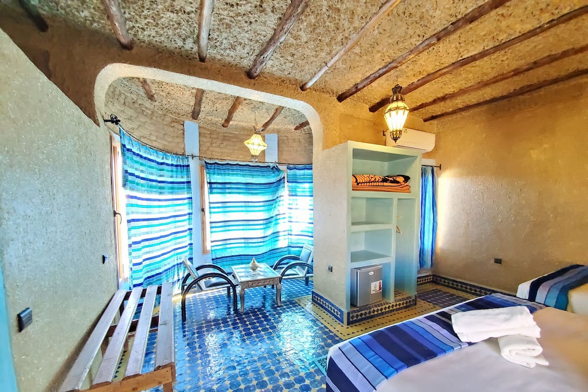A room with a tiled floor, a bed, a desk with a chair, a wardrobe, and a small refrigerator.