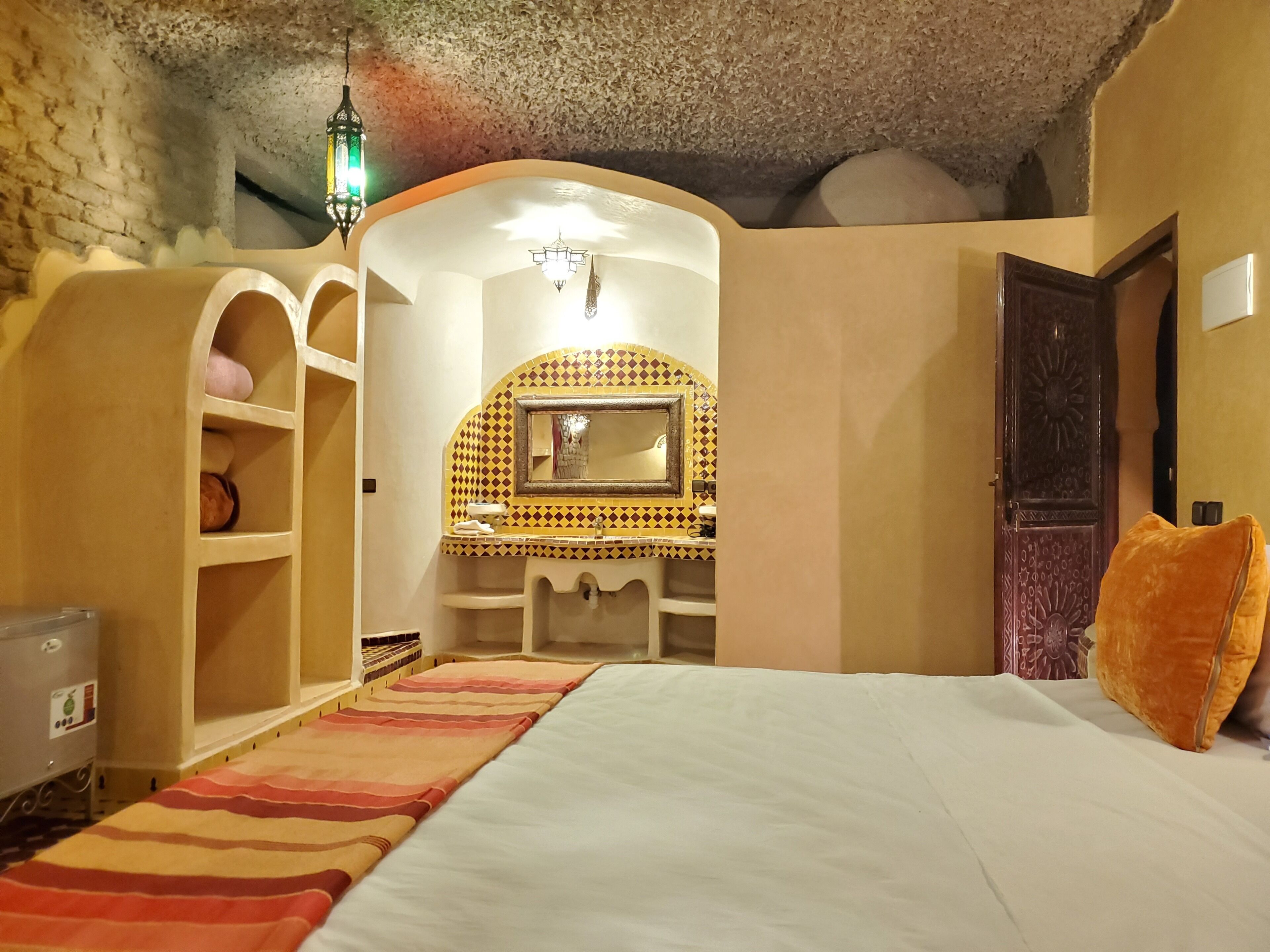 Une chambre à coucher comprenant un lit, une coiffeuse avec un miroir, des étagères, une lanterne suspendue et une porte ornée de motifs complexes.