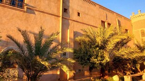 A building with a flat facade, multiple windows, and a balcony. There are palm trees and a paved area in front. - Ksar Merzouga  (Taouz)