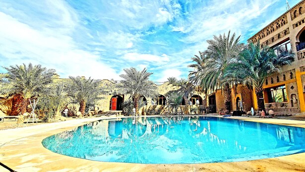 A swimming pool with a curved edge, surrounded by palm trees and a building with arches and balconies. - Ksar Merzouga  (Taouz)