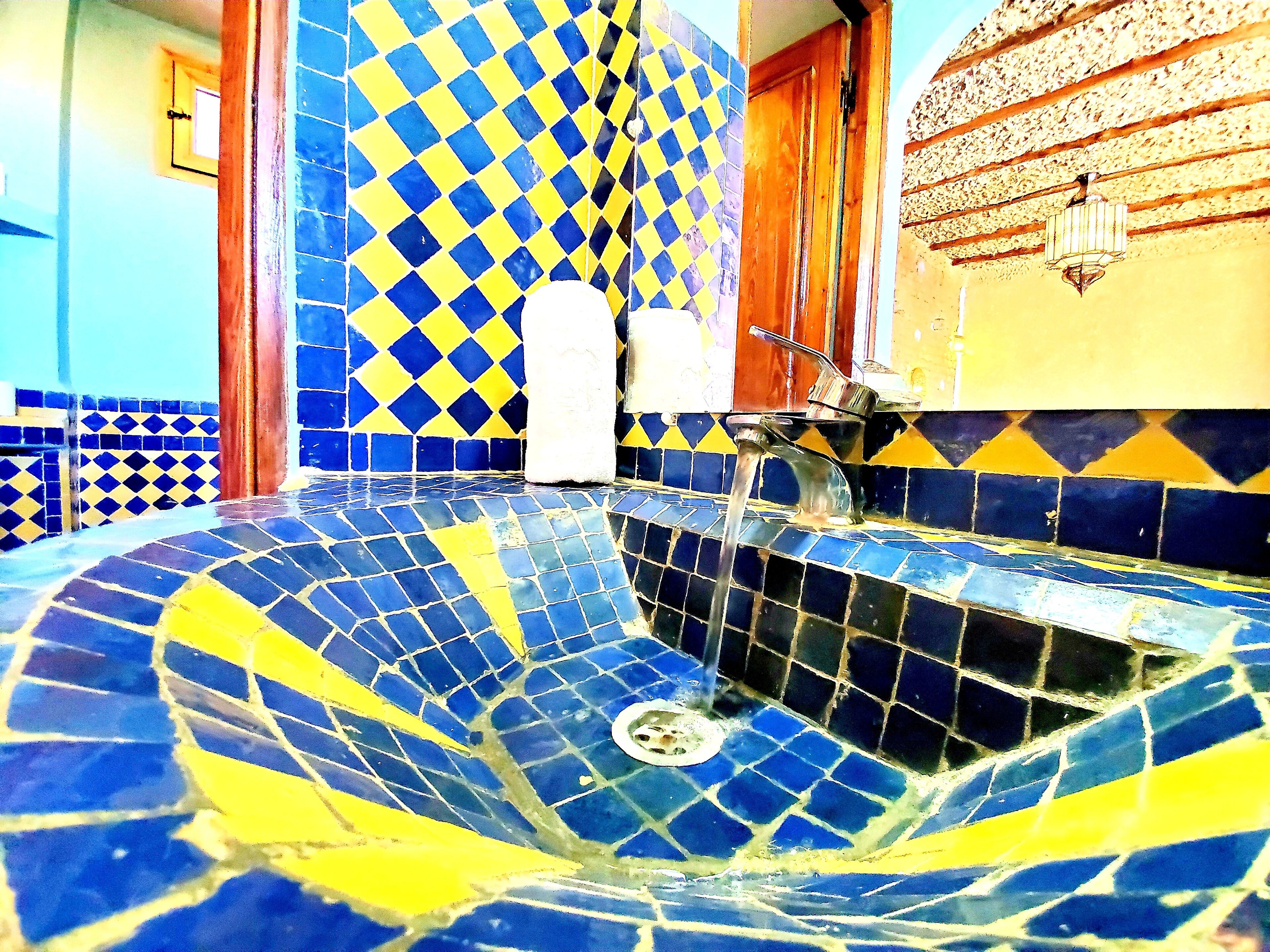 Een badkamer met een blauw en geel betegelde vloer, een witte zuil en een houten deur.