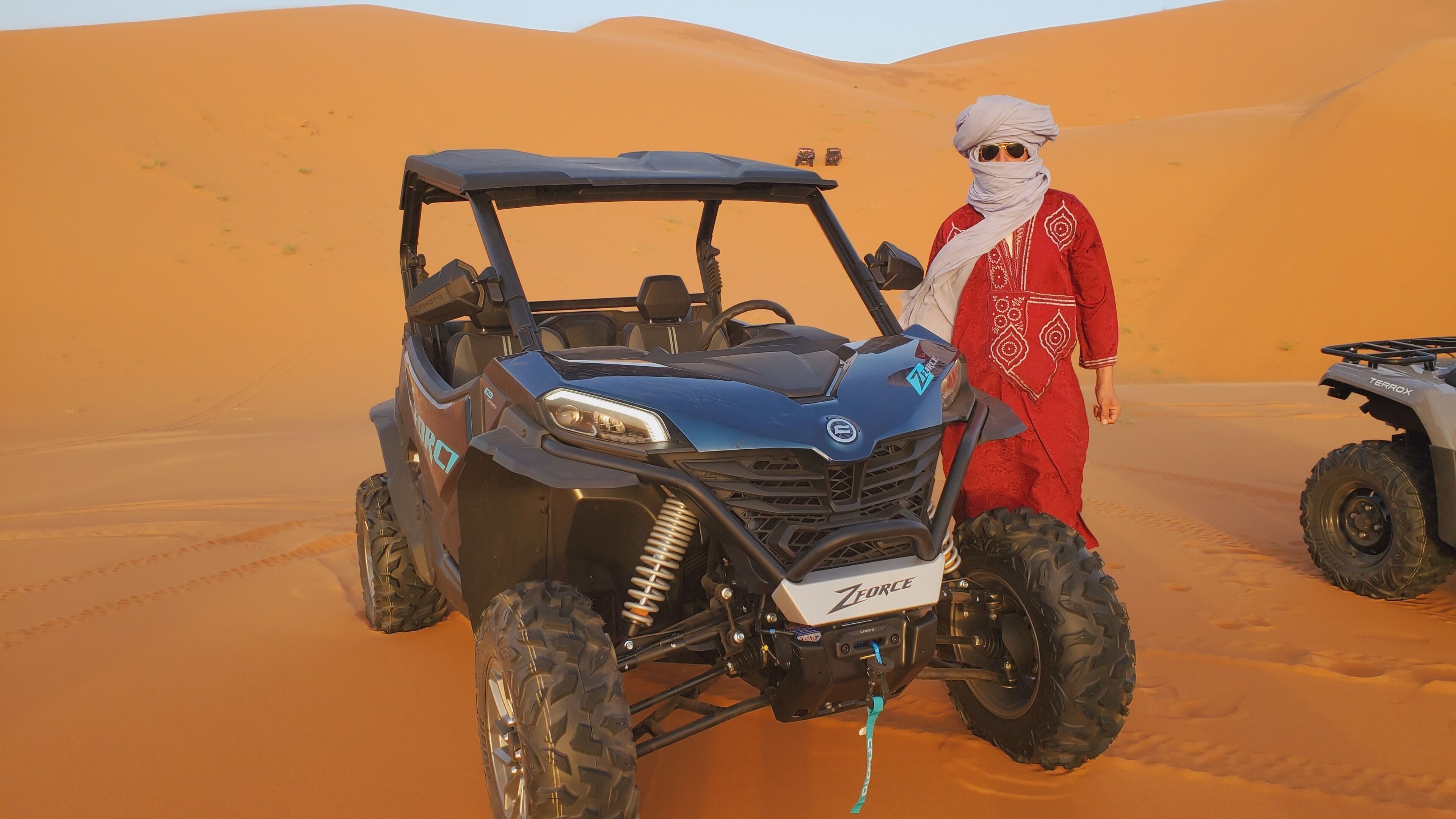 A desert scene with a blue off-road vehicle and a person in traditional attire standing beside it.