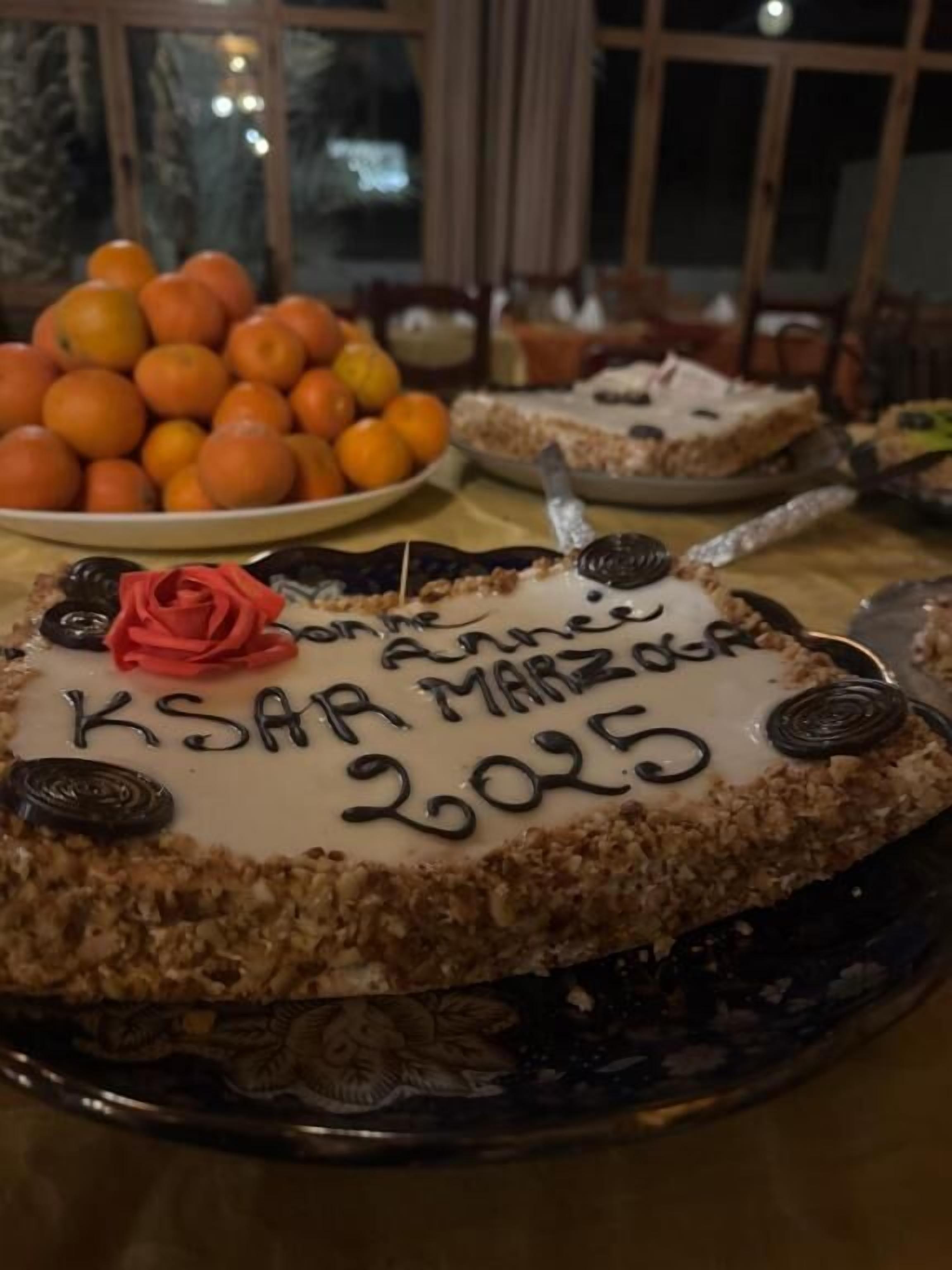 A cake with "KSAAR MARZOOG 2025" written on it, surrounded by oranges and other desserts on a table.