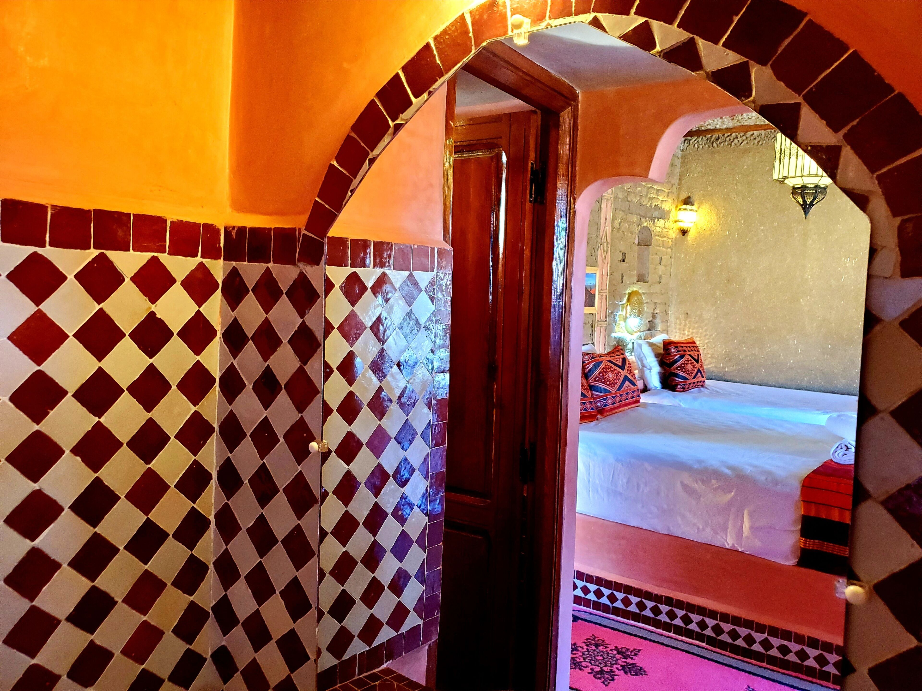 Una habitación con piso a cuadros, una cama con almohadas estampadas y un espejo que refleja una cama y una pared con una lámpara decorativa.