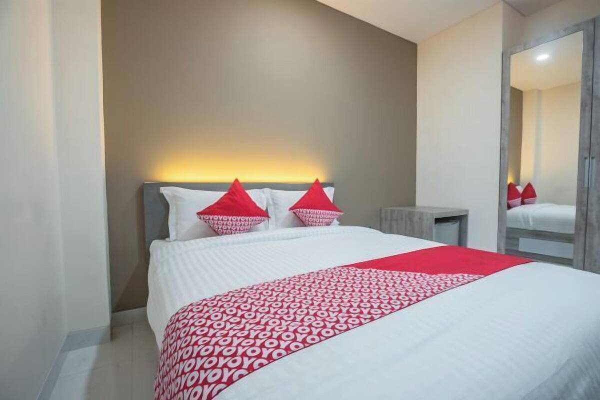Una cama bien hecha con una colcha de cuadros rojos y blancos, un cabecero con dos almohadas rojas y una mesita de noche con una lámpara.