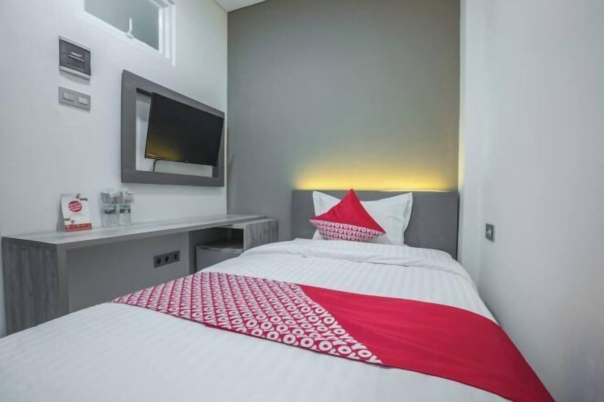 Habitación de hotel con cama, televisor montado en la pared, mesita de noche y una manta decorativa con estampado rojo y blanco.