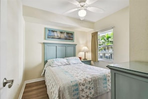 Une chambre à coucher avec un lit, une table de chevet, une lampe, un ventilateur de plafond, une fenêtre avec des rideaux et un tableau au mur.