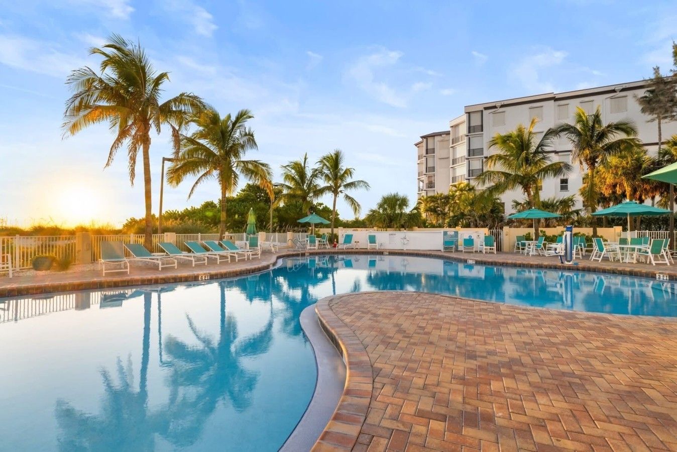 A swimming pool with lounge chairs and palm trees, surrounded by a building with multiple floors.