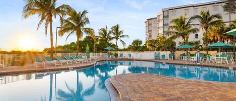 A swimming pool with lounge chairs and palm trees, surrounded by a building with multiple floors.