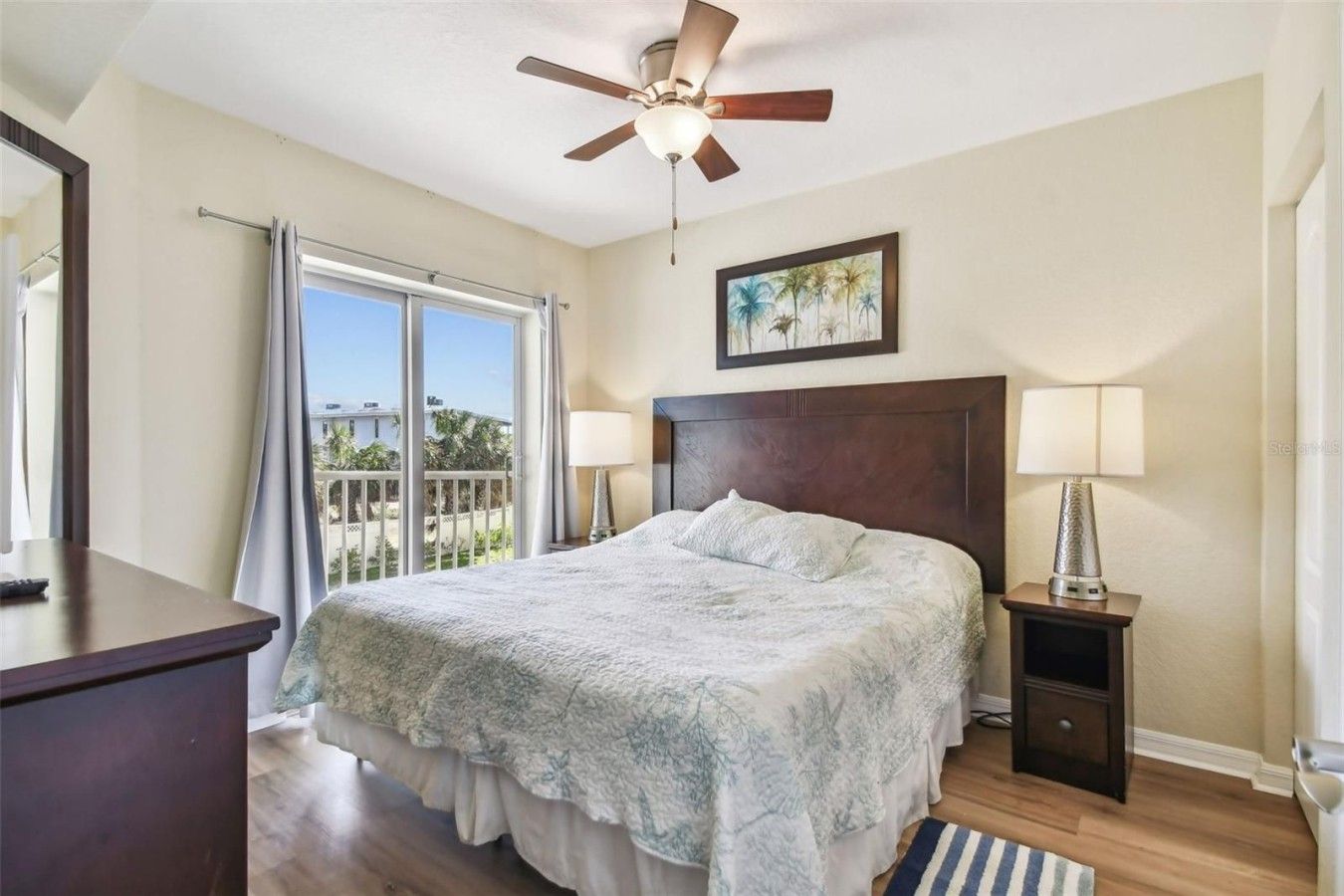 A bedroom with a bed, a ceiling fan, a nightstand with a lamp, a window with curtains, and a painting on the wall.