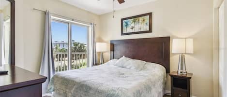 A bedroom with a bed, a ceiling fan, a nightstand with a lamp, a window with curtains, and a painting on the wall.