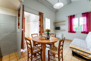 A room with a round wooden dining table, four chairs, a sofa, a TV on a shelf, and a window with red curtains. - Holiday Home 'Gite Fontenelle Alpilles' with Shared Pool, Wi-Fi & Air Conditioning (Arles)