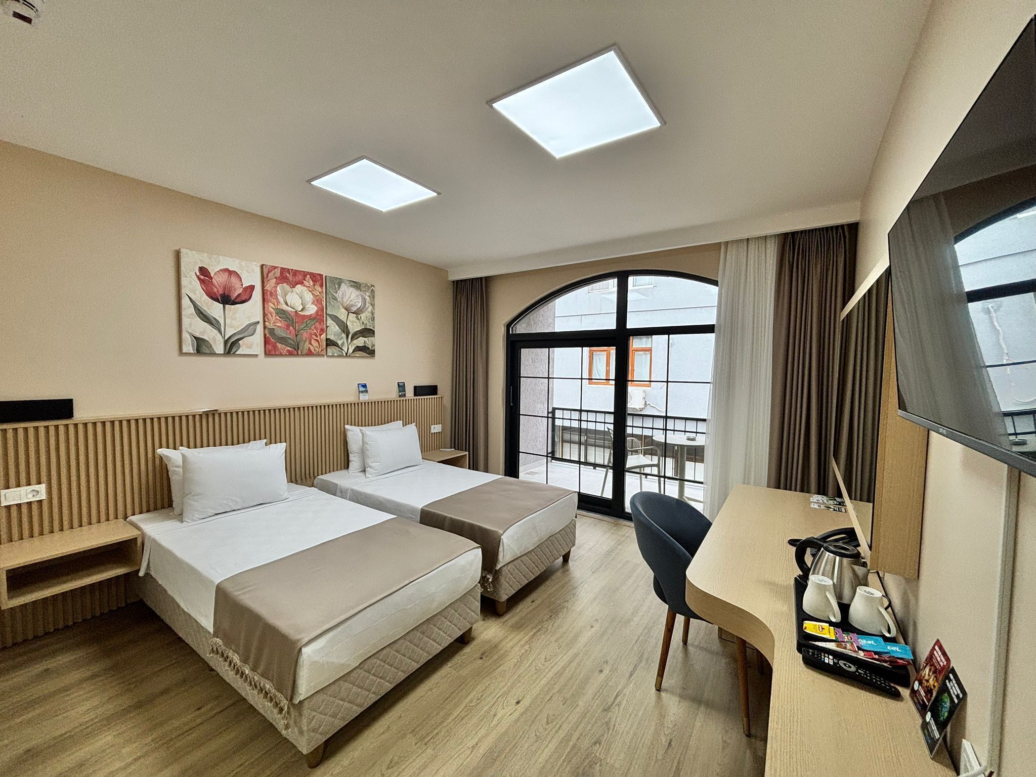 A hotel room with two beds, a desk with a chair, a large window with curtains, and artwork on the wall.