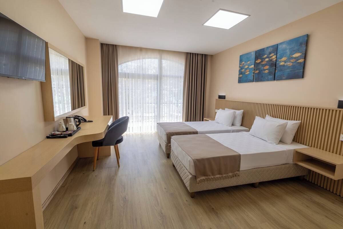 A hotel room with two beds, a desk, a chair, a TV, and a large window with curtains.