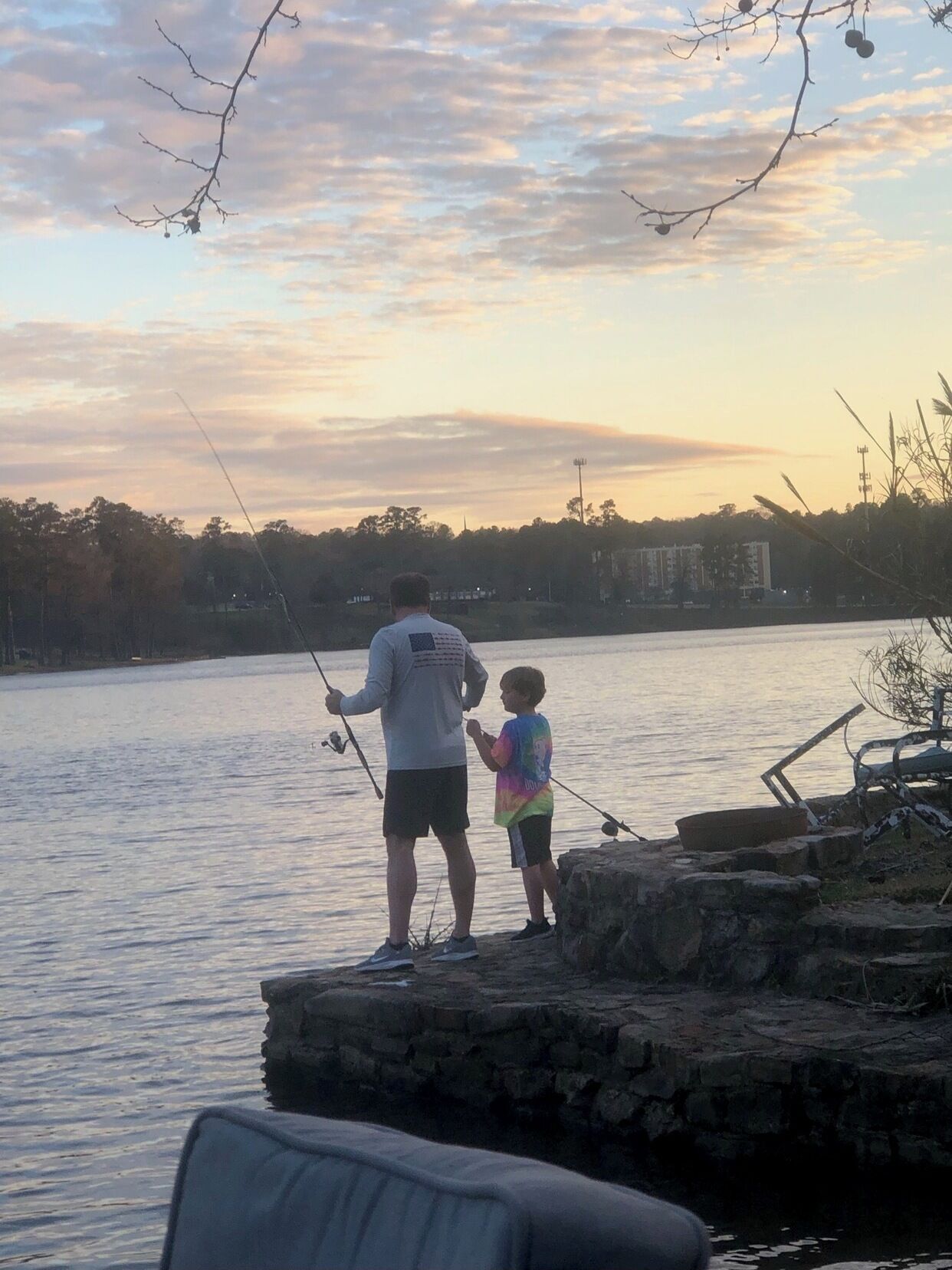 Un hombre y un niño pescan en un lago tranquilo con un muelle de piedra y una silla en primer plano.