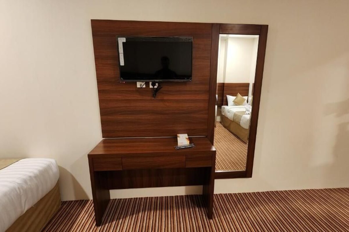 A hotel room with a wooden desk, a flat-screen TV mounted on the wall, a bed, and a mirror reflecting the room.