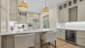Una cocina moderna con armarios blancos, una isla central con taburetes y iluminación colgante, además de vistas a zonas verdes.