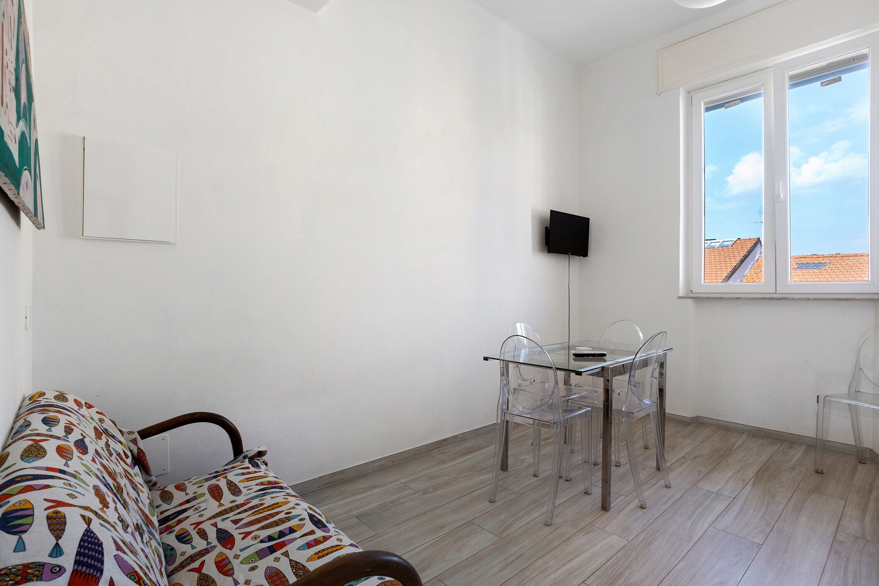 Quarto com sofá, mesa de jantar com cadeiras, TV fixada na parede e janela com vista para o céu.