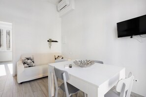 A modern, white interior with a dining table, chairs, a sofa, and a flat-screen TV mounted on the wall.