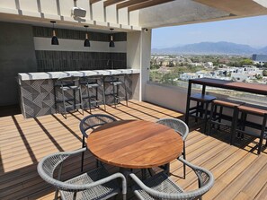 Eine Holzterrasse mit einem runden Tisch und Stühlen, eine Bar mit Hockern und Blick auf eine Stadtlandschaft mit Bergen im Hintergrund.