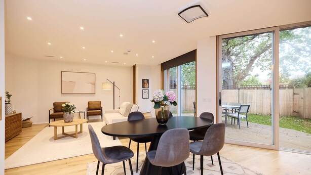 Een ruime woonkamer met een ronde zwarte tafel, grijze stoelen, een witte bank en een grote glazen schuindeur naar een buitenruimte.