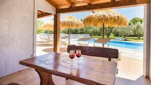 A wooden table with two glasses of red drink, a wooden bench, and a view of a swimming pool with lounge chairs and thatched umbrellas.