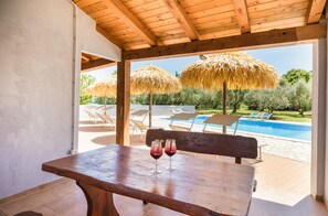 Una mesa de madera con dos vasos de bebida roja, un banco de madera y vistas a una piscina con tumbonas y sombrillas de paja.